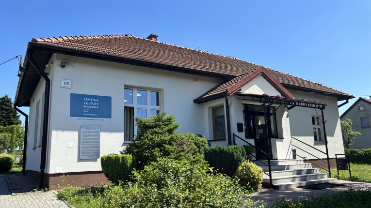 budynek klubu Soboniowice, na środku wejście przez ganek po kilku schodach, po lewej stronie powieszona flaga Polski, duże okna, czterospadzisty dach, chodnik prowadzący do wejście, zieleń dookoła budynku