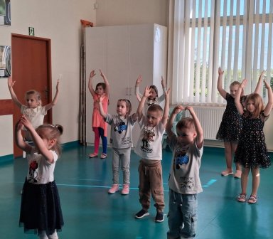 Tańczące dzieci na sali w klubie kultury z rękami nad głową, kolorowe stroje.